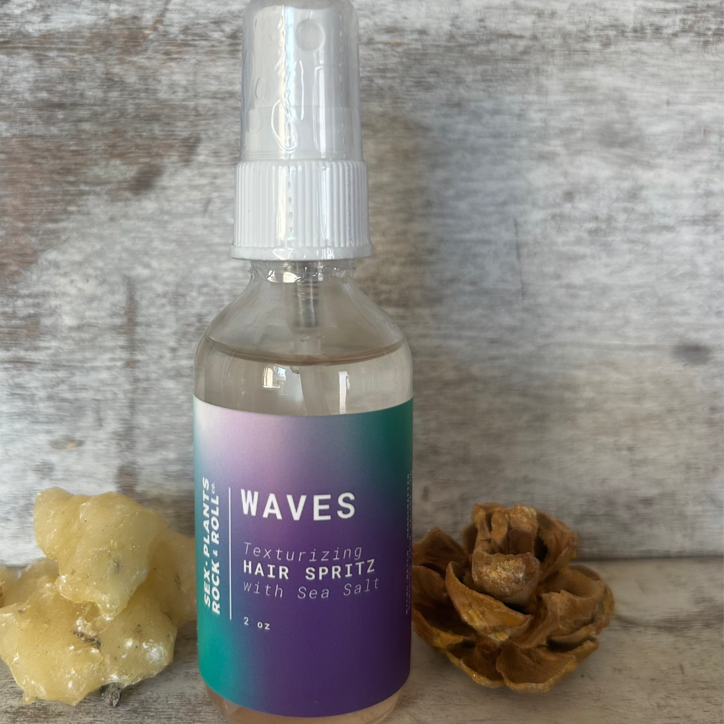 A bottle of 'Waves - Texturizing Hair Spritz with Sea Salt' positioned on a wooden surface, with a pine cone and what appears to be a piece of driftwood in the background, suggesting an ocean-inspired theme.