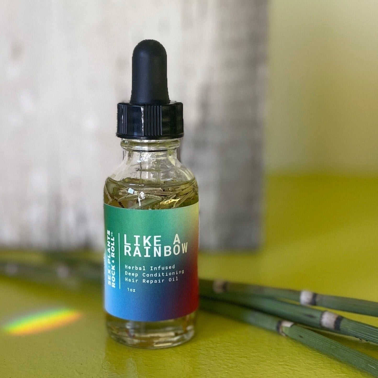 small clear glass dropper bottle with oil and herbs rainbow label and white lettering reads like a rainbow herbal infused deep conditioning hair repair oil