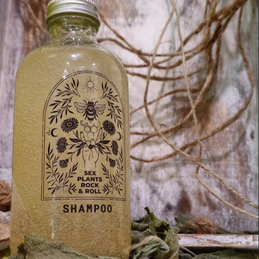 glass bottle with golden bubble shampoo in it with a clear label, black design, rock and roll hand holding calendula flowers, a bee with a honeycomb below it, the sun on top and two crescent moon designs on either side with herbs filling in the space