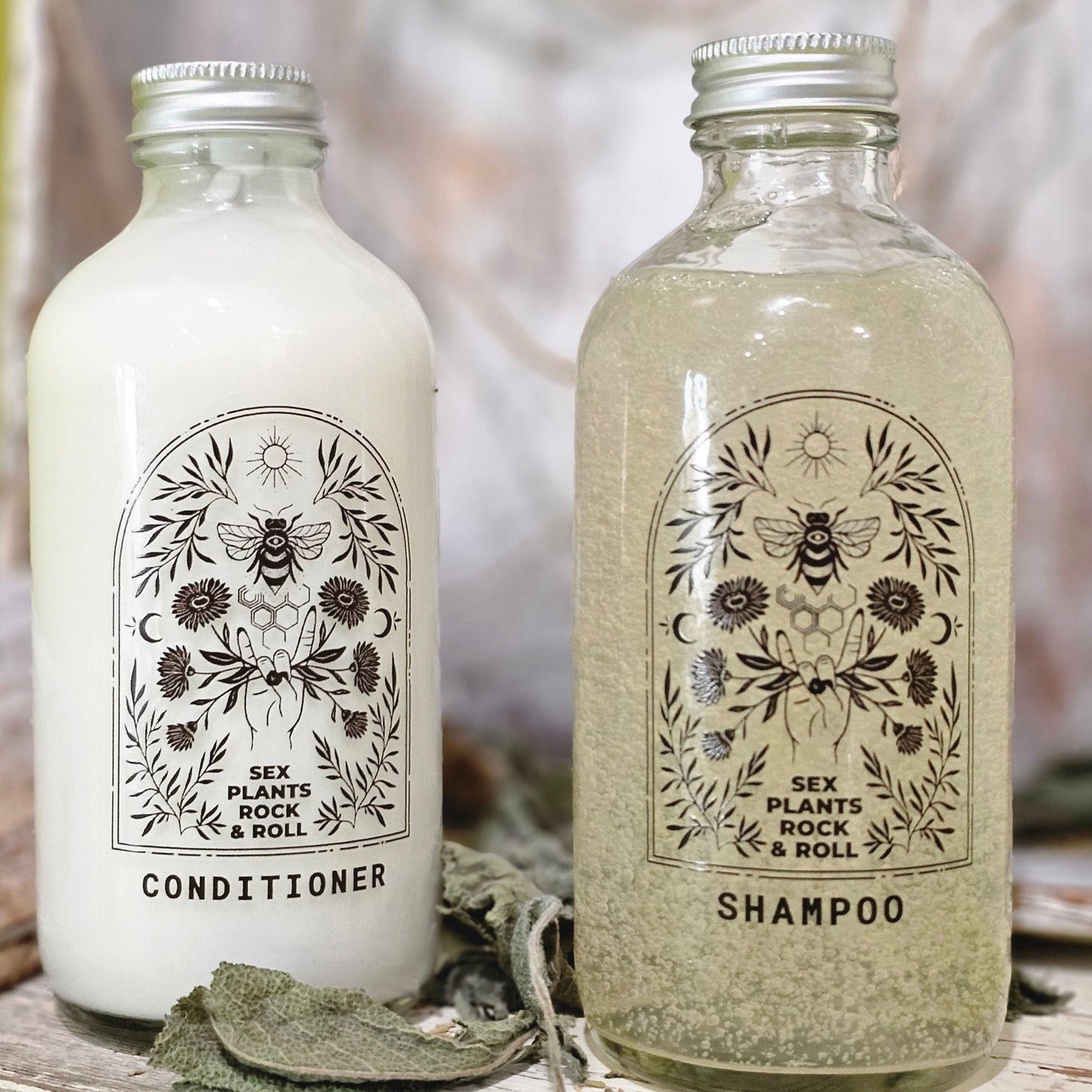 two glass bottles. one labeled conditioner with white conditioner in it. one labeled shampoo with bubley shampoo in it. Both have a clear label, black design, rock and roll hand holding calendula flowers, a bee with a honeycomb below it, the sun on top and two crescent moon designs on either side with herbs filling in the space