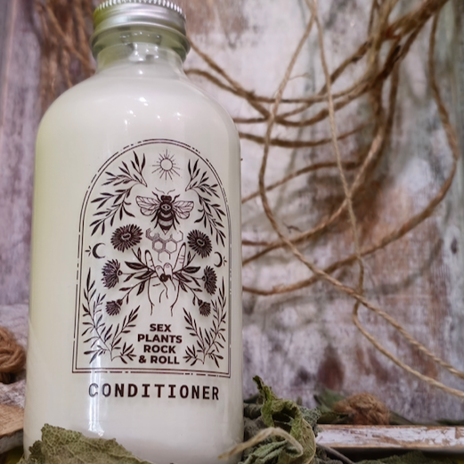 glass bottle with white conditioner in it with a clear label, black design, rock and roll hand holding calendula flowers, a bee with a honeycomb below it, the sun on top and two crescent moon designs on either side with herbs filling in the space